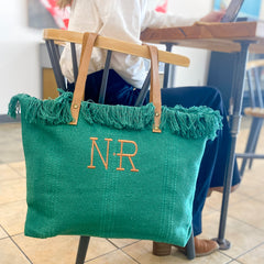 The emerald green tote bag for women is hanging from the back of a chair at a table in a restaurant. The personalized tote has two modern font letters with a dot in between them in a camel thread that coordinates with the vegan leather handles. 