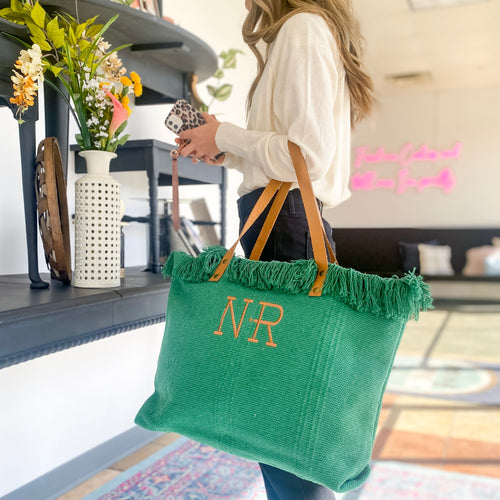 A model is standing, looking at a display, with an emerald green fringe tote on her arm. The vegan leather straps are at her elbow and are attached to the bag at the top. The body of the bag is textured and woven in a cotton material.