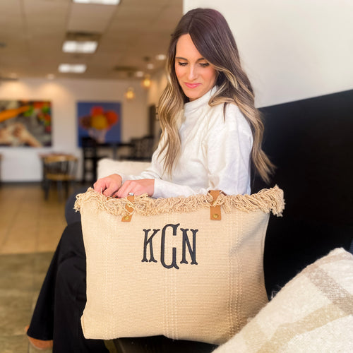 Khaki colored, textured tote bag sits in a bakery on a black bench. The top of the bag has fringe detailing. A model is unzipping the closure of the bag at the top. The vegan leather handles are camel colored and folded back.