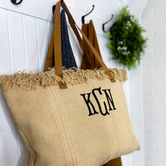 The Khaki utility tote is hanging on a white hall tree with sweaters and a wreath in the background. The monogrammed tote has large, embroidered letters in the upper center of the bag. It is personalized with black thread and a serif font. 