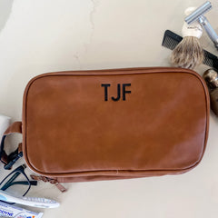 Brown, rectangular travel kit laying flat on a white counter. Matching handle on the left side shows a black buckle that can be used to attach it to a larger bag. Various toiletry items surround the bag.