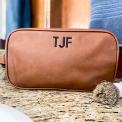 Brown, vegan leather dopp kit sitting on a granite countertop. Handle on the left side. Staged with a towel behind the bag, and a shaving brush in front of it. The bag is embroidered at the top with small initials in black thread and block font.