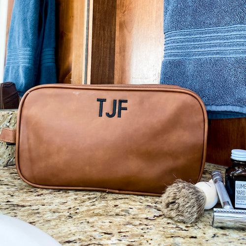 Brown, vegan leather dopp kit sitting on a granite countertop. Handle on the left side. Staged with a towel behind the bag, and a shaving brush and razor in front of it. The bag is embroidered at the top with small initials in black thread and block font.