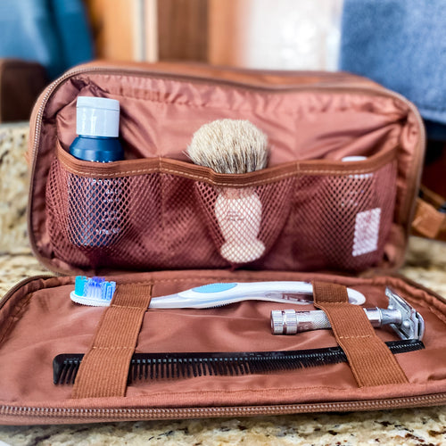 Outer dopp kit flap is zipped open and lined. Front panel lays flat with elastic bands, holding toothbrush, razor and comb. Panel on the body of the bag has 3 open pockets holding a bottle of beard oil, a shaving brush, and an after shave bottle. 