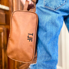 Brown travel case is being held by the vegan leather handle in a vertical direction by a model dressed in denim jeans. Brown vegan leather is smooth and varigated in coloring for a vintage look. Travel kit is personalized with same thread and font. 