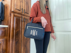 Black hanging travel case in a bathroom setting. Model has makeup bag through her arm and the top handles rest at her elbows. 3 inch monogram and zippered pocket shown on the front panel. Model is in a rust sweatshirt and black leggings.