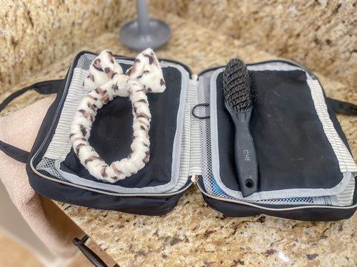Toiletry case is laid on a granite bathroom counter over a towel. Sections are folded inward. Gold zipper hardware will close the main section of the kit. Additional items like brushes and headbands can be stowed in this compartment.