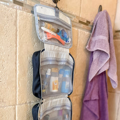 Toiletry bag is hanging by the attached metal hook in a travertine shower on a hook on the wall. All compartments are folded out vertically. Sections are filled with various toiletries and makeup. There is a set of towels on the other hook.