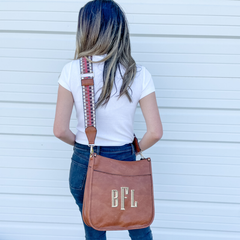 Woman modeling a crossbody guitar strap purse. The purse is brown with a weaved strap that has brown, rust, white, and sliver of hunter green. Purse features monogram initials in tan.