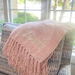 Blush colored chenille blanket is folded and laying on the edge of Chair. Blanket has fluffy fringe and an embroidered name in a fun font with greige thread.