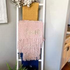 Blush colored chenille blanket is folded and hanging on a white blanket ladder Blanket has fluffy fringe and an embroidered name in a fun font with greige thread.