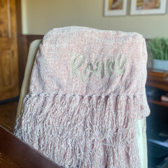 Blush colored chenille blanket is folded and draped on office chair.  Blanket has fluffy fringe and an embroidered name in a fun font with greige thread.