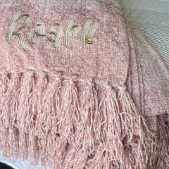 Blush colored chenille blanket is folded and laying on the edge of a bed with white linens. Blanket has fluffy fringe and an embroidered name in a fun font with greige thread.