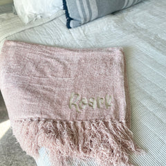 Blush colored chenille blanket is folded and laying on the edge of a bed with white linens. Blanket has fluffy fringe and an embroidered name in a fun font with greige thread.