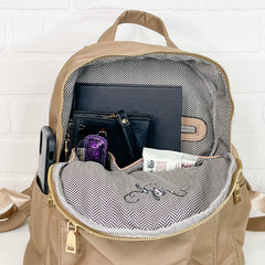 Tan backpack is open holding a book and wallet in main compartment. in front of a back zipper. Two small open pockets holding glasses and mascara and other a snack bar. Side Exterior pocket holding phone.