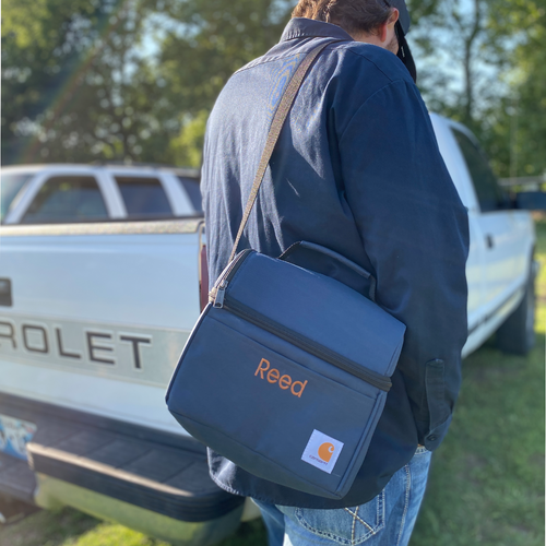 A Navy Carhartt lunch box slung over the shoulder of a model. Cooler has a handle with a dome-shaped top. Silver hardware. Front pocket on lower, squared section is adorned with embroidered name in camel thread. Carhartt logo on bottom right corner.