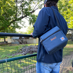 A Navy Carhartt lunch box slung over the shoulder of a model. Cooler has a handle with a dome-shaped top. Silver hardware. Front pocket on lower, squared section is adorned with embroidered name in camel thread. Carhartt logo on bottom right corner.