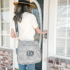 Woman showing a light grey crossbody purse. Purse features a floral patttern guitar strap with a black base and white and light pink embroidered flowers. Large script monogram in black is displayed on purse.