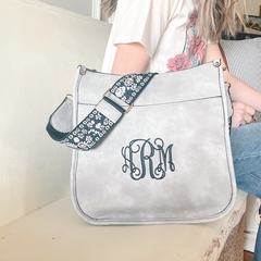 Light grey guitar strap purse sitting on a bench next to a lady. Purse has black strap with floral light pink and white embroidery. Purse has large black monogram initials.