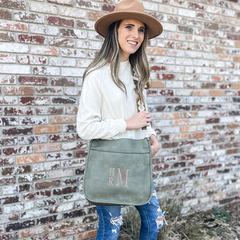 Woman standing in front of a brick wall wearing a hat and sweater. Modeling a Guitar strap crossbody purse. The crossbody is olive green and strap features a unique design. 